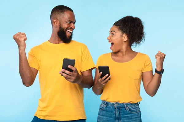 Online win, shopping sale concept. Excited millennial black couple using mobile phones, gesturing YES on blue background — Fotografia de Stock