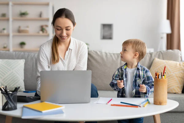 Adorable toddler boy drawing next to mom working on laptop, lady spending time with son and enjoying freelance work — Fotografia de Stock