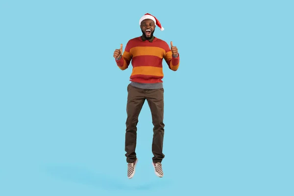 Tatil Teklifi. Noel Baba Şapkalı Siyah Adam Zıplıyor ve Baş Parmakla İşaret Ediyor — Stok fotoğraf