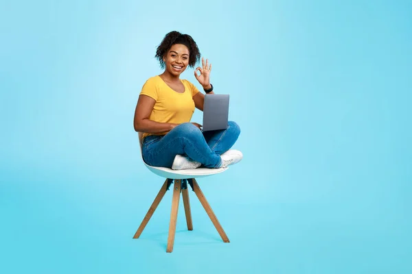 若い黒の女性は、ラップトップで椅子に座って、良いジェスチャーを示し、オンライン仕事や教育をお勧めします — ストック写真