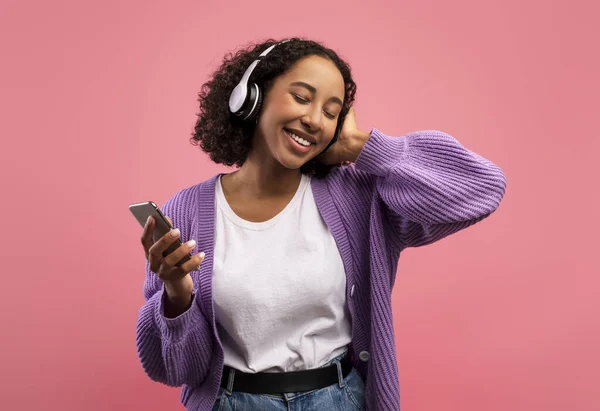 Positive young black woman with headphones and cellphone listening to music, using new mobile app on pink background — Foto Stock