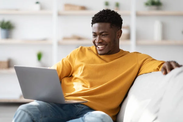 Chico negro alegre sentado en el sofá con el ordenador portátil — Foto de Stock