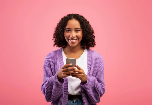 Cheery young black lady holding modern smartphone, working remotely or video chatting on pink studio background — Foto de Stock