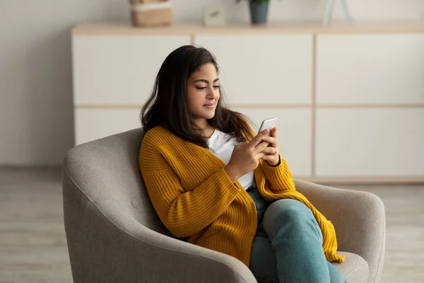 Check social networks at home. Positive arab woman using cellphone app, typing message, relaxing in armchair —  Fotos de Stock