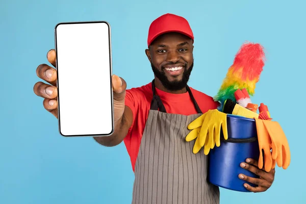 Black male cleaner holding basket with cleaning tools and showing blank smartphone — Fotografia de Stock