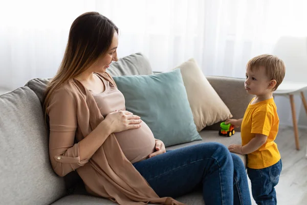 Young pregnant mother playing with her son, sitting on sofa and talking to her little child while spending time together — 스톡 사진