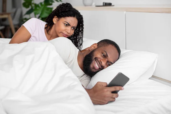 Upset millennial black female looks at smartphone of her husband, chatting on phone in social networks and play — 스톡 사진