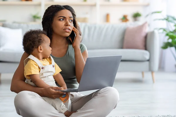 Black Mom Using Laptop And Talking On Phone With Baby On Hands — стоковое фото