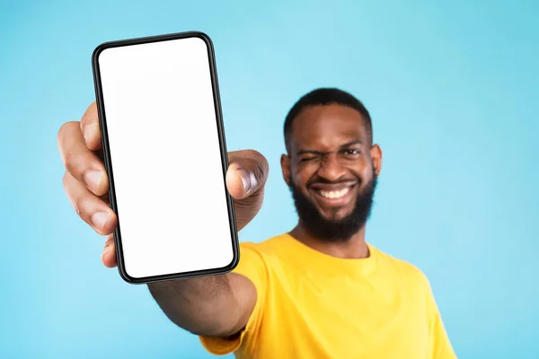 Mobile app advertisement. Smiling black man showing smartphone with empty screen, winking on blue background, mockup — Stockfoto