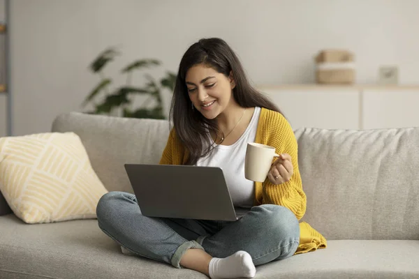 Morning concept. Excited arab female freelancer drinking coffee at home and using laptop, working online, free space — Fotografia de Stock