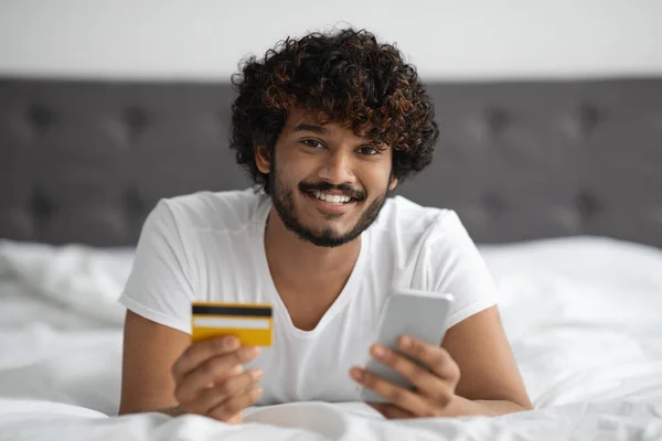 Chico feliz acostado en la cama con tarjeta de crédito y teléfono celular —  Fotos de Stock