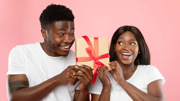 Feliz pareja negra sacudiendo caja de regalo en el estudio rosa —  Fotos de Stock