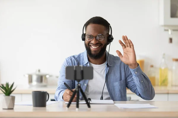 Smiling young black male blogger with beard in glasses waving hand, looks at phone, working remotely — Foto Stock