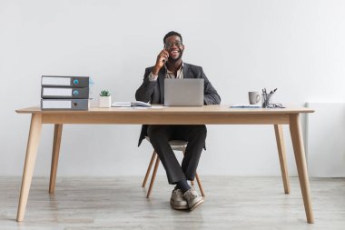 Cheerful black businessman in formal suit talking on smartphone, having conversation, sitting at table with laptop