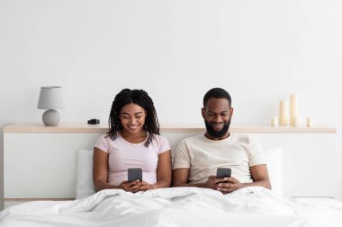 Smiling busy young black female and male sitting on bed and typing on smartphones in bedroom interior