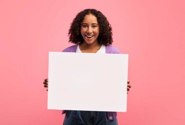 Happy millennial black woman holding empty poster, smiling at camera on pink studio background, mockup