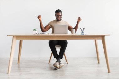 Successful black man making YES gesture while working on laptop at desk against white studio wall, copy space