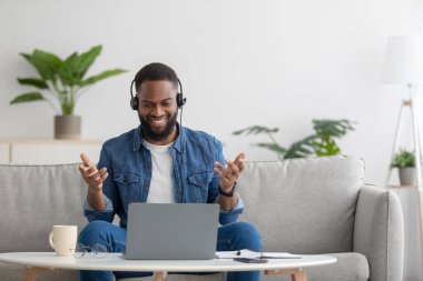 Cheerful glad young black businessman, teacher with beard in headphones with pc gesturing with hands
