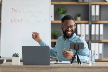 Smiling handsome young black male tutor with beard show rules of english and looks at pc and smartphone