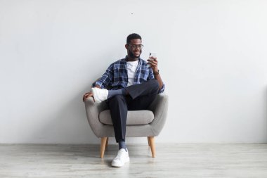 Happy black guy sitting in armchair with smartphone, texting to friend, sending message, browsing web against white wall