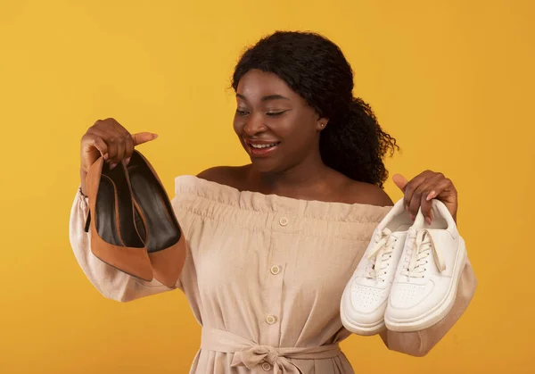 Young African American female selecting between high-heeled shoes and comfortable sneakers on orange background — Fotografia de Stock
