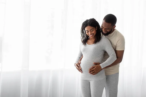 Satisfied millennial black handsome hugs pregnant wife in bedroom interior and look at belly — стоковое фото
