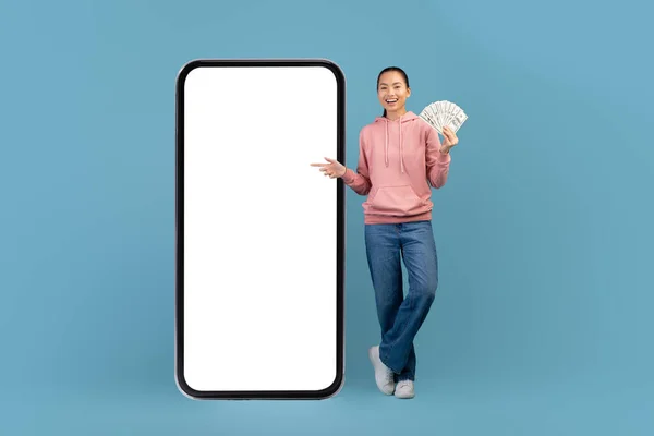 Cheerful asian woman with bunch of dollars and big smartphone — 图库照片