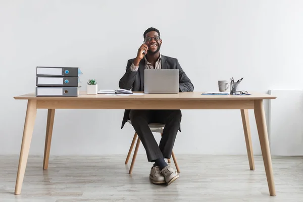 Веселий чорний бізнесмен в офіційному костюмі розмовляє на смартфоні, розмовляє, сидить за столом з ноутбуком — стокове фото