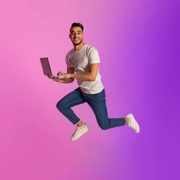 Full length of millennial Arab man with laptop computer jumping up in neon light. Cool new website or advertisement — Fotografia de Stock