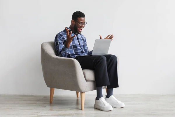 Young black man gesturing YES, sitting in armchair with laptop, celebrating success or achievement against white wall — Fotografia de Stock