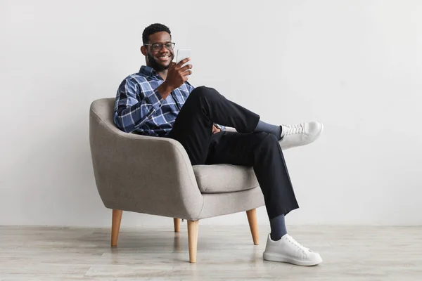 Joyful young black guy sitting in armchair with smartphone, working remotely or video chatting against white wall — Stockfoto