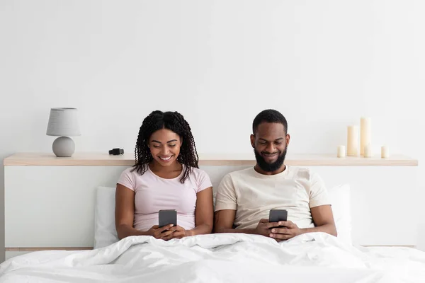 Smiling busy young black female and male sitting on bed and typing on smartphones in bedroom interior — Stock fotografie