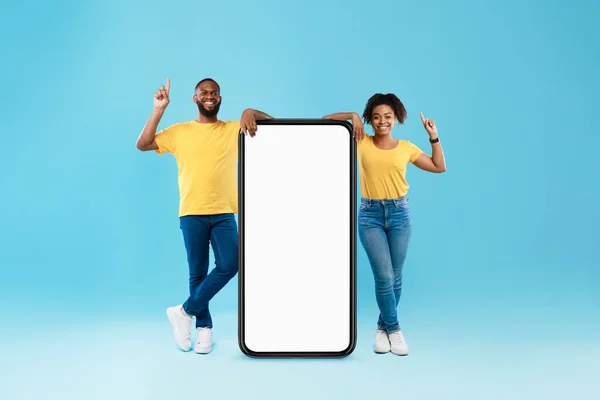 Positive black couple demonstrating huge smartphone, offering mockup for your app or websiteon empty screen, pointing up — 图库照片