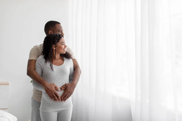Cheerful relaxed millennial black guy hugs lady in bedroom interior and look out window — Stock fotografie