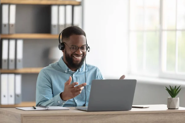 Cheerful attractive millennial black male with beard in glasses, headphones gestures with hands look at pc — Stok fotoğraf