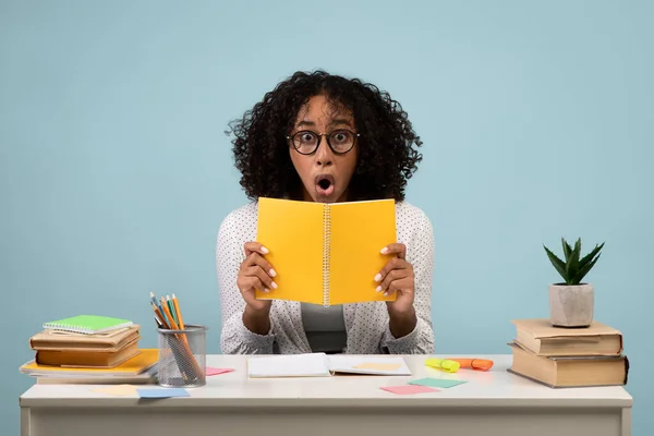 Portrait of shocked young black woman in eyeglasses reading notebook, sitting at table on blue studio background — Stock Photo, Image