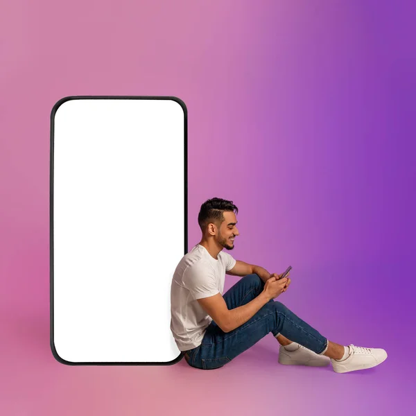 Chico árabe alegre sentado cerca de un gran teléfono celular con pantalla en blanco, utilizando el dispositivo móvil, utilizando la nueva aplicación en luz de neón, maqueta —  Fotos de Stock