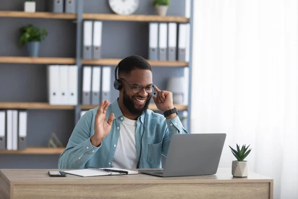 Amistoso joven empresario afroamericano feliz con barba en gafas y auriculares funciona en PC y agita la mano — Foto de Stock