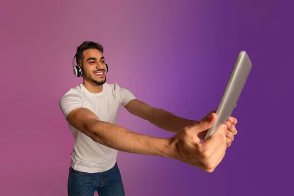 Handsome millennial Arab man chatting online, using tablet pc, wearing wireless headphones in neon light — Fotografia de Stock