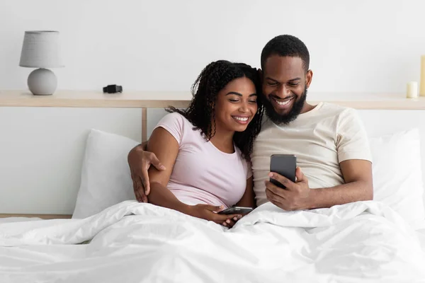 Cheerful young black lady and guy sitting on bed hugging and looking at phone, watch video — 스톡 사진