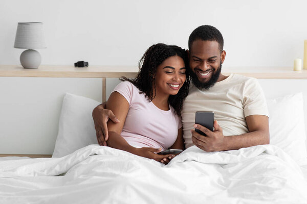 Cheerful young black lady and guy sitting on bed hugging and looking at phone, watch video