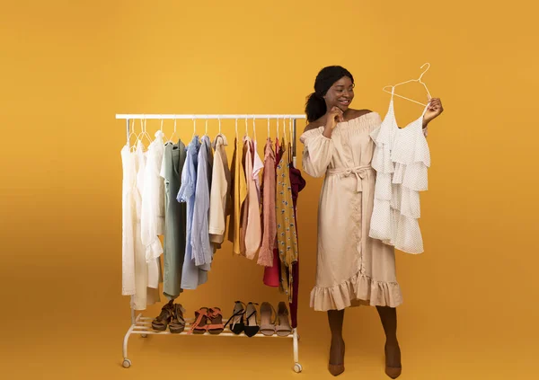 Black female stylist holding dress near clothing rack, choosing trendy outfit on orange background. Shopping concept — Fotografia de Stock