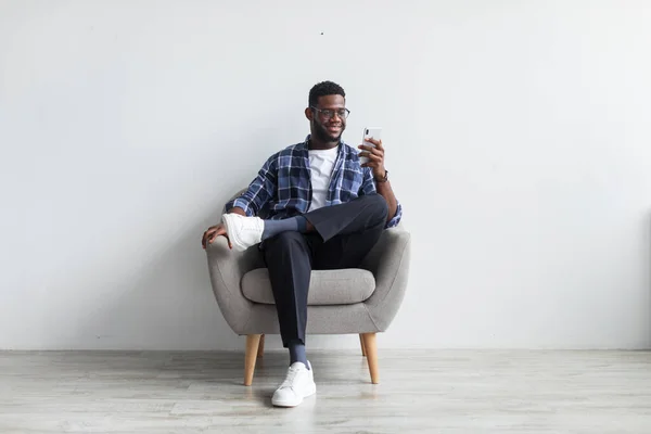 Happy black guy sitting in armchair with smartphone, texting to friend, sending message, browsing web against white wall — Foto de Stock