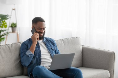 Busy cheerful millennial african american male with beard looks at pc, call to client by phone in living room interior