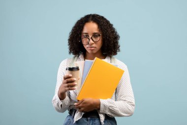 Tired young black female student with notebooks and takeout coffee looking at camera on blue studio background