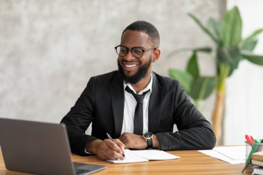 African American man in glasses using laptop writing in notebook