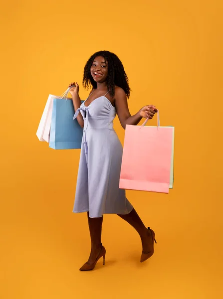 Happy young black woman in pretty dress holding bright shopping bags, walking on orange studio background — Fotografia de Stock