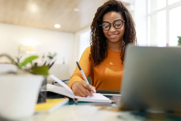 Frau sitzt am Schreibtisch, benutzt Computer und schreibt in Notizbuch — Stockfoto