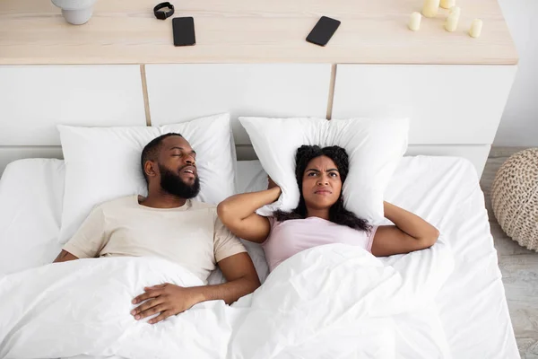 Upset despair millennial black woman covers ears with pillow, suffers from snoring of husband on bed — 스톡 사진