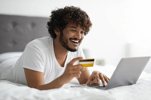 Chico feliz acostado en la cama con tarjeta de crédito y portátil —  Fotos de Stock